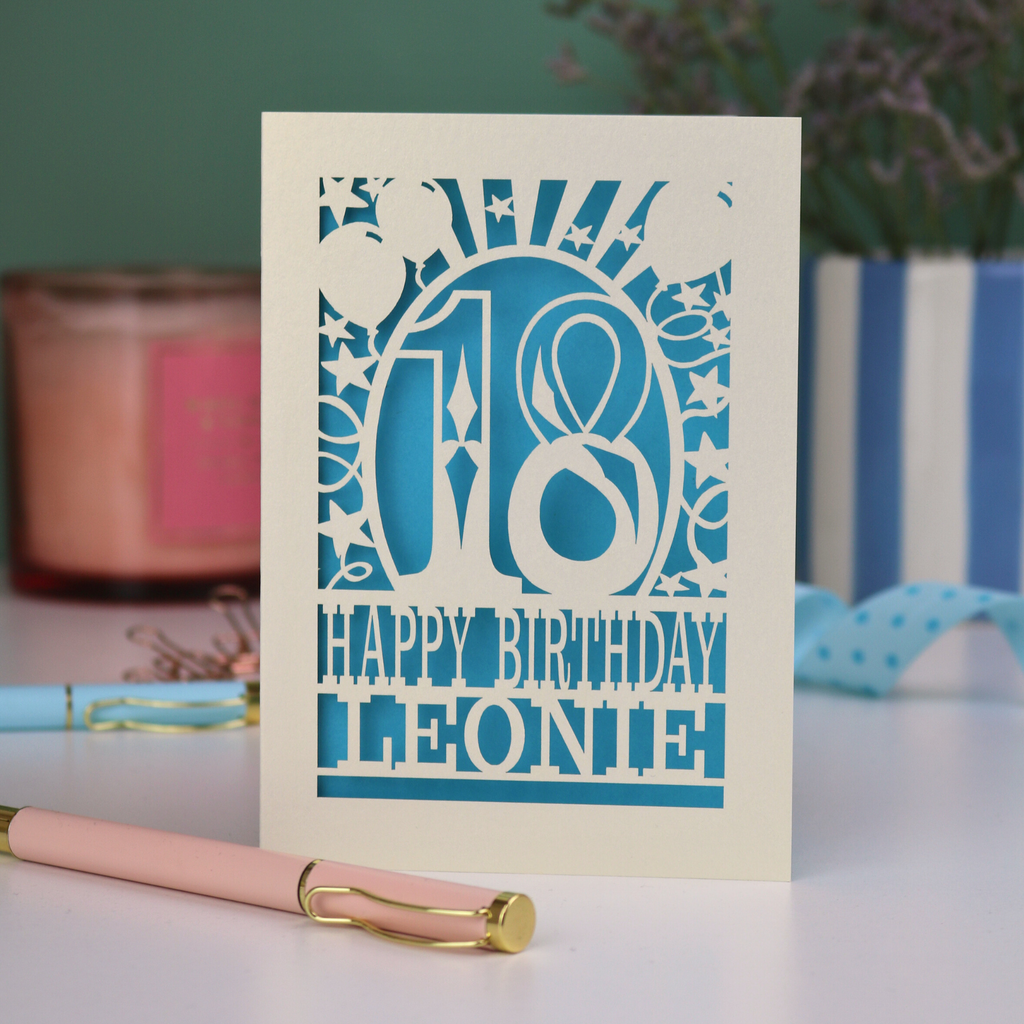A personalised laser cut 18th birthday card with 18, Happy Birthday, Name. Card is cut out from a cream card with a bright blue paper insert.