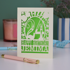 A cream laser cut birthday card on a white desk with stationery items. Card has the age 24 surrounded by stars and balloons, with "Happy Birthday Jemima" underneath in cut out text