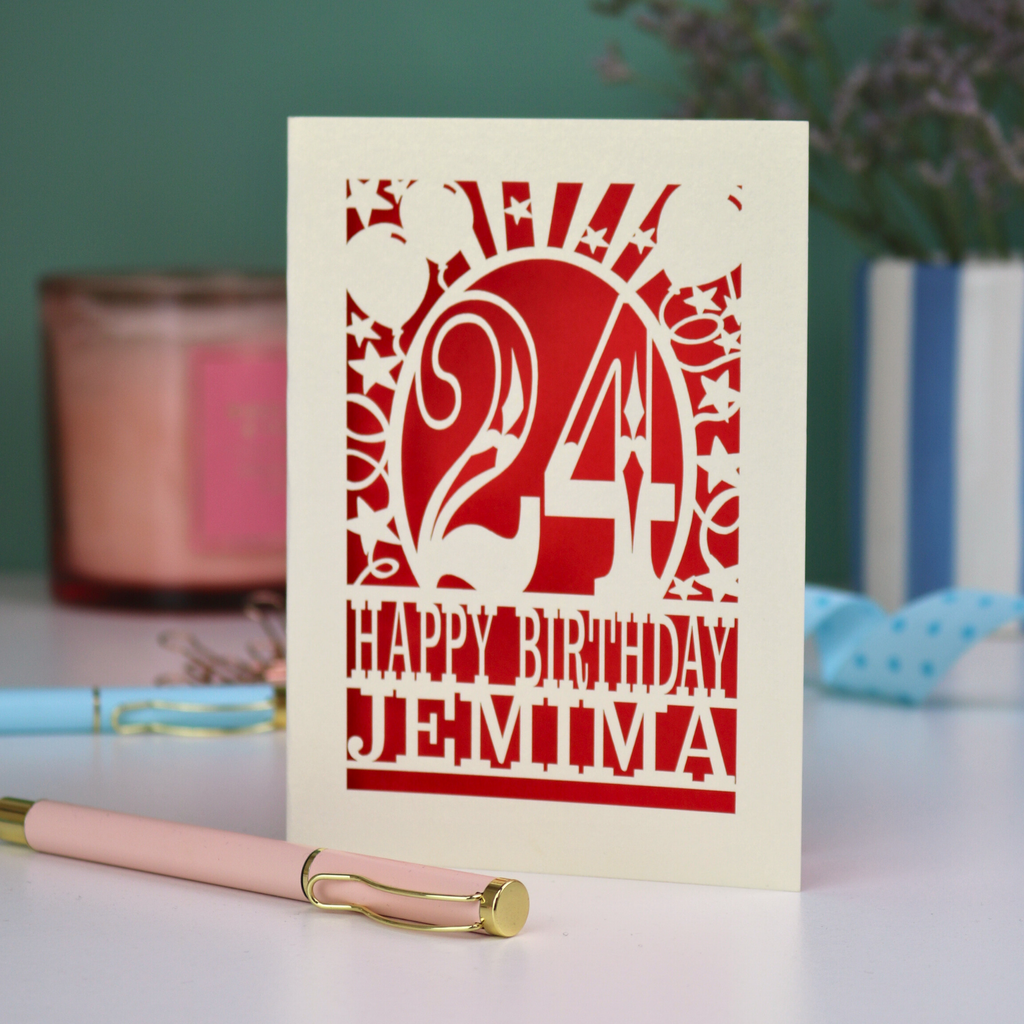 A cream laser cut birthday card on a white desk with stationery items. Card has the age 24 surrounded by stars and balloons, with "Happy Birthday Jemima" underneath in cut out text