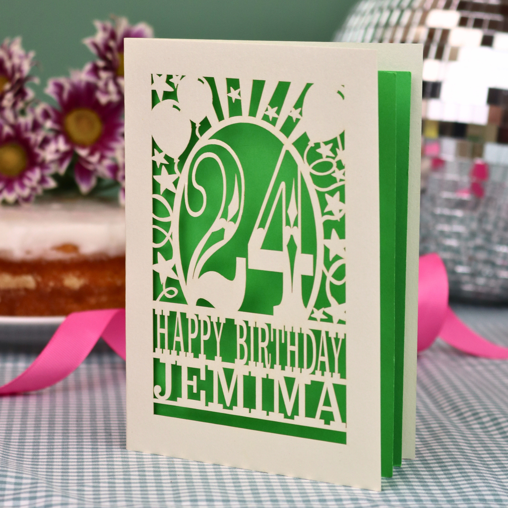 A personalised birthday card with a laser-cut design, featuring a cream-coloured cardstock with a bright green cut out design with the number 24 and the text 'HAPPY BIRTHDAY JEMIMA'. The card is standing upright on a checked tablecloth with pink ribbons and blurred background elements.