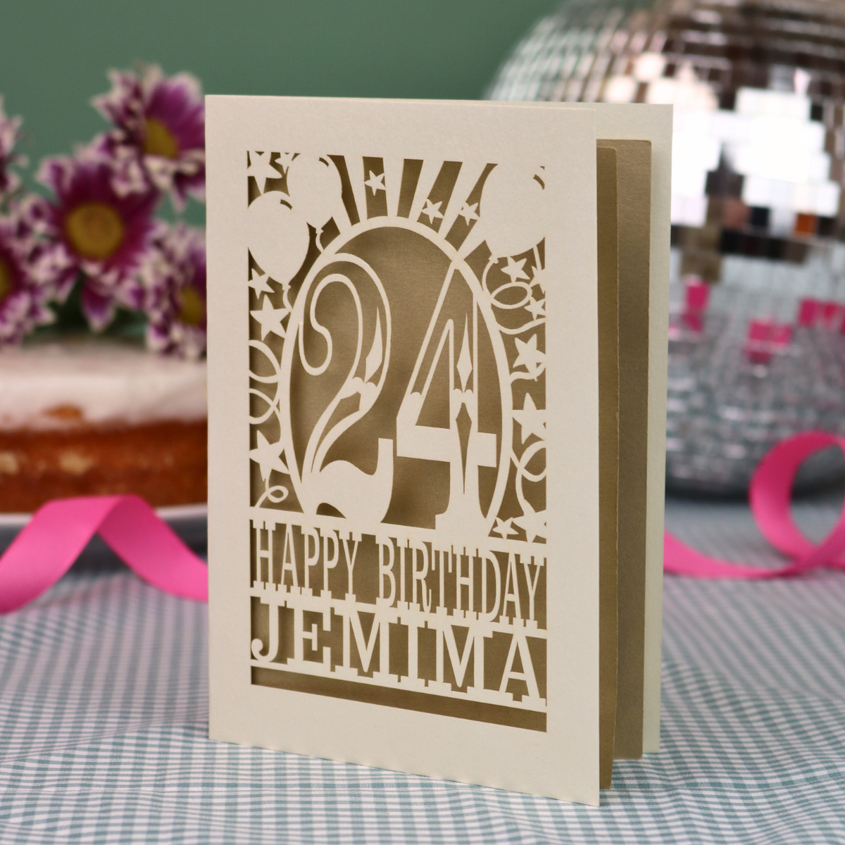 A personalised birthday card with a laser-cut design, featuring a cream-coloured cardstock with a gold leaf cut out design with the number 24 and the text 'HAPPY BIRTHDAY JEMIMA'. The card is standing upright on a checked tablecloth with pink ribbons and blurred background elements.