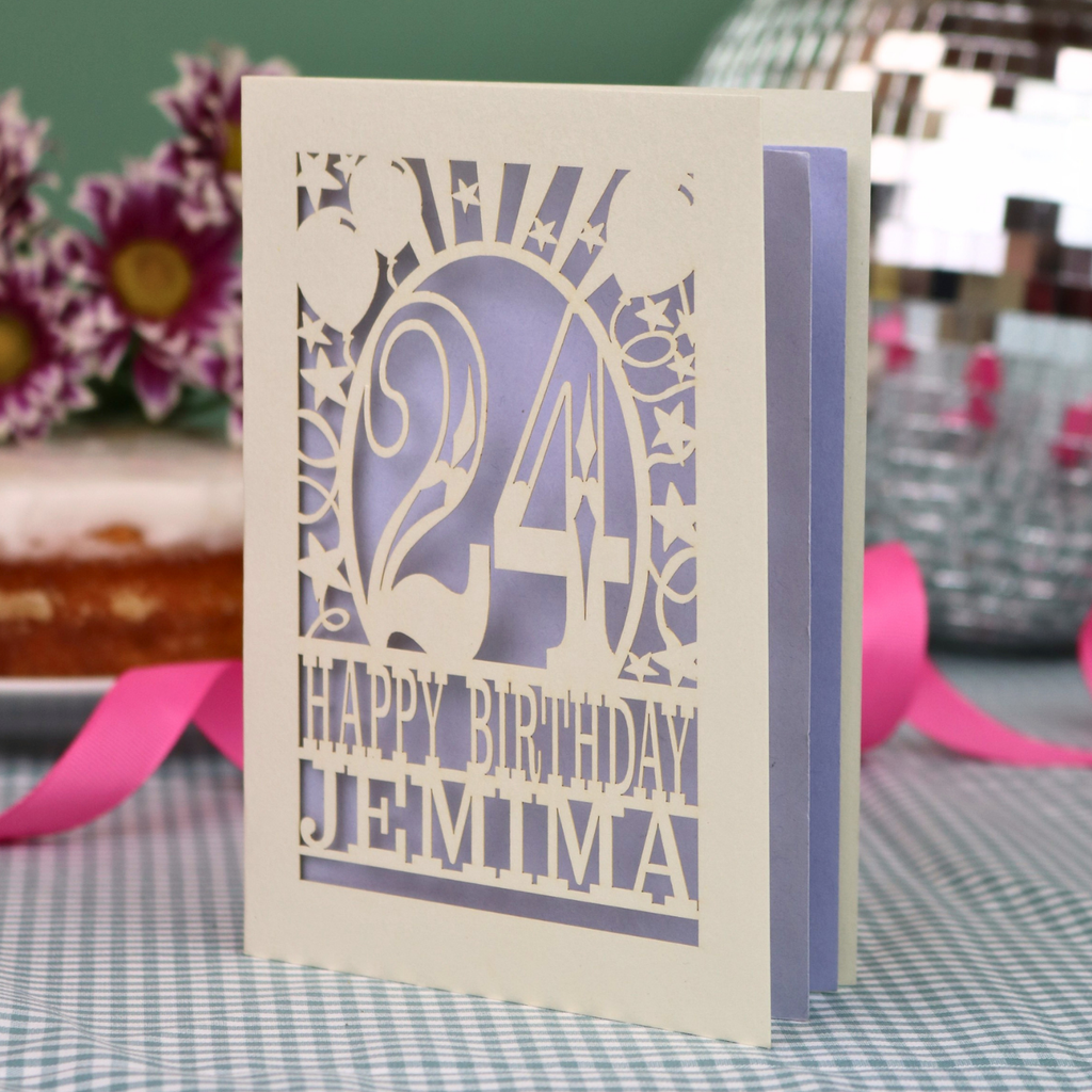 A personalised birthday card with a laser-cut design, featuring a cream-coloured cardstock with a lilac paper insert and cut out design with the number 24 and the text 'HAPPY BIRTHDAY JEMIMA'. The card is standing upright on a checked tablecloth with pink ribbons and blurred background elements.