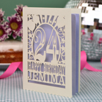 A personalised birthday card with a laser-cut design, featuring a cream-coloured cardstock with a lilac paper insert and cut out design with the number 24 and the text 'HAPPY BIRTHDAY JEMIMA'. The card is standing upright on a checked tablecloth with pink ribbons and blurred background elements.