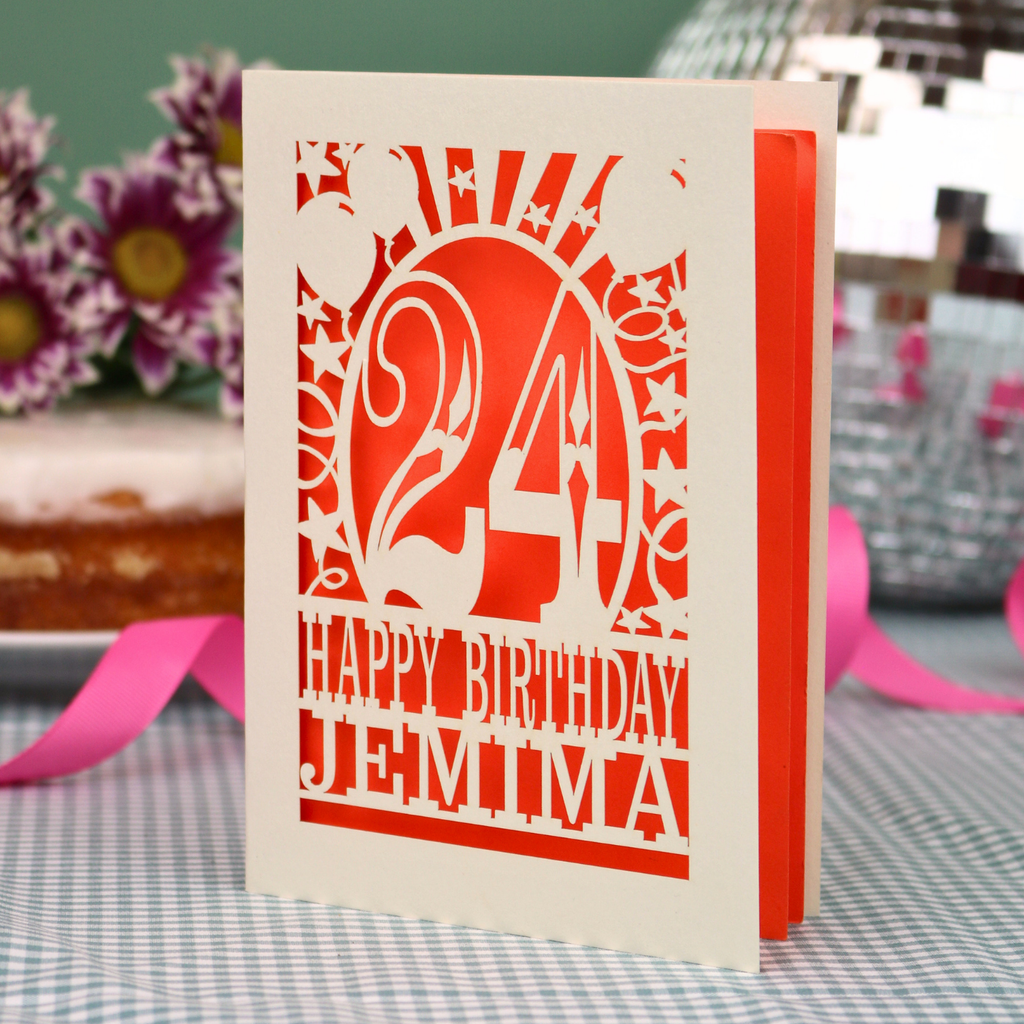 A personalised birthday card with a laser-cut design, featuring a cream-coloured cardstock with orange paper insert and cut out design with the number 24 and the text 'HAPPY BIRTHDAY JEMIMA'. The card is standing upright on a checked tablecloth with pink ribbons and blurred background elements.