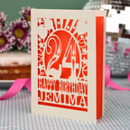 A personalised birthday card with a laser-cut design, featuring a cream-coloured cardstock with orange paper insert and cut out design with the number 24 and the text 'HAPPY BIRTHDAY JEMIMA'. The card is standing upright on a checked tablecloth with pink ribbons and blurred background elements.