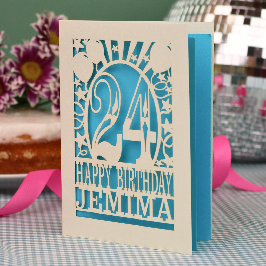 A personalised birthday card with a laser-cut design, featuring a cream-coloured cardstock with a blue paper insert and cut out design with the number 24 and the text 'HAPPY BIRTHDAY JEMIMA'. The card is standing upright on a checked tablecloth with pink ribbons and blurred background elements.
