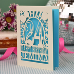 A personalised birthday card with a laser-cut design, featuring a cream-coloured cardstock with a blue paper insert and cut out design with the number 24 and the text 'HAPPY BIRTHDAY JEMIMA'. The card is standing upright on a checked tablecloth with pink ribbons and blurred background elements.