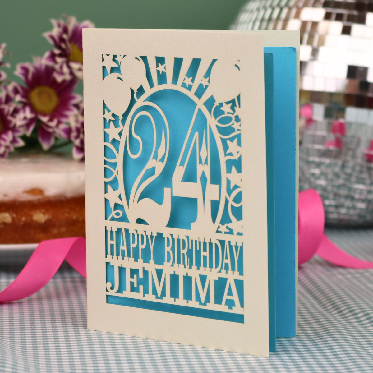 A personalised birthday card with a laser-cut design, featuring a cream-coloured cardstock with a blue paper insert and cut out design with the number 24 and the text 'HAPPY BIRTHDAY JEMIMA'. The card is standing upright on a checked tablecloth with pink ribbons and blurred background elements.