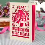 A personalised birthday card with a laser-cut design, featuring a cream-coloured cardstock with a hot pink cut out design with the number 24 and the text 'HAPPY BIRTHDAY JEMIMA'. The card is standing upright on a checked tablecloth with pink ribbons and blurred background elements.