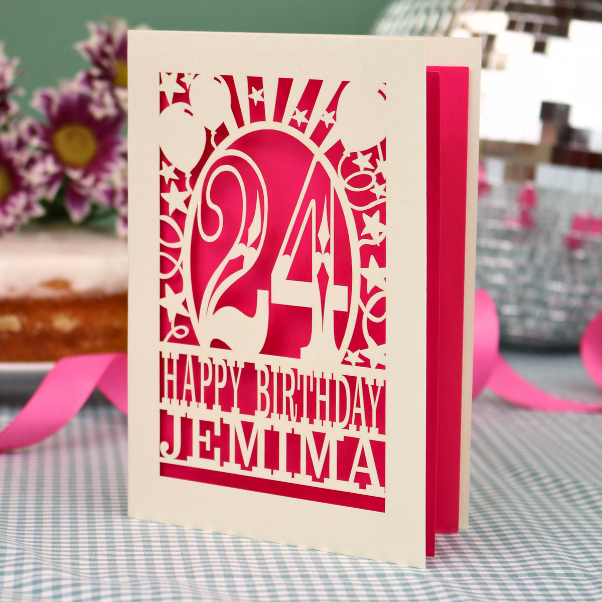 A personalised birthday card with a laser-cut design, featuring a cream-coloured cardstock with a hot pink cut out design with the number 24 and the text 'HAPPY BIRTHDAY JEMIMA'. The card is standing upright on a checked tablecloth with pink ribbons and blurred background elements.