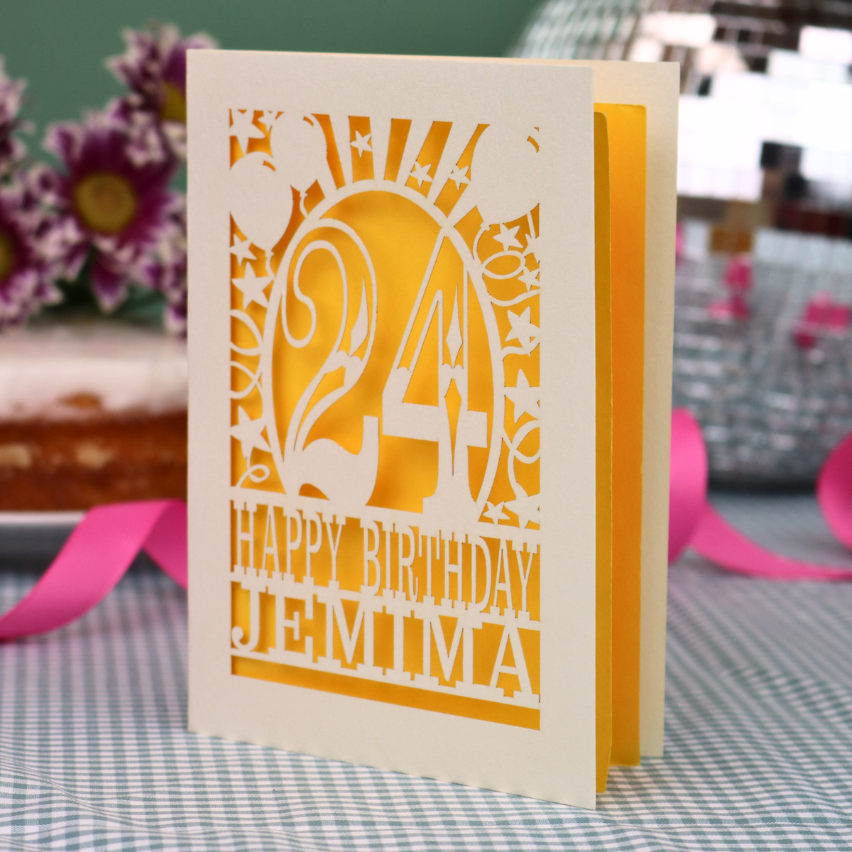 A personalised birthday card with a laser-cut design, featuring a cream-coloured cardstock with a bright yellow paper insert and cut out design with the number 24 and the text 'HAPPY BIRTHDAY JEMIMA'. The card is standing upright on a checked tablecloth with pink ribbons and blurred background elements.