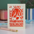 A cream and orange laser cut birthday card on a white desk with stationery items. Card has the age 24 surrounded by stars and balloons, with "Happy Birthday Jemima" underneath in cut out text