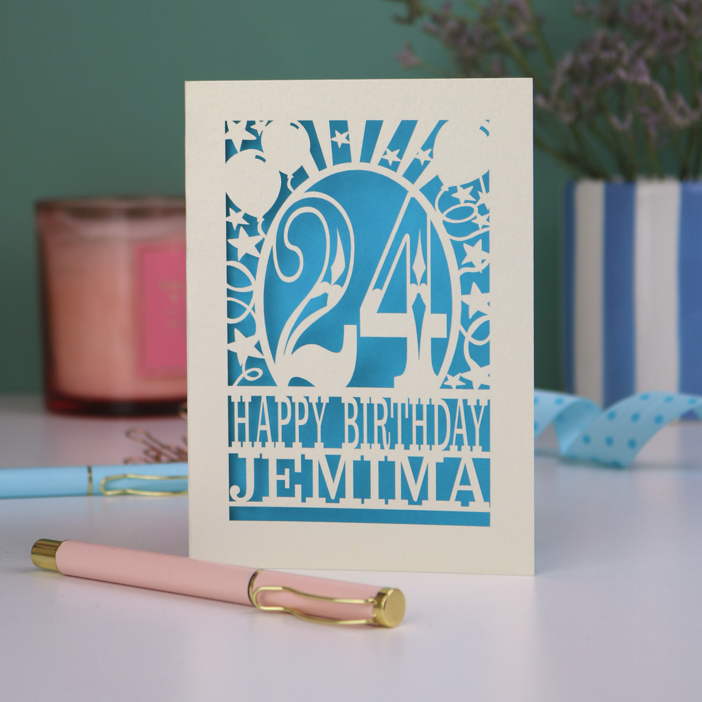 A cream personalised birthday card on a white desk with stationery items. Card has the age 24 surrounded by stars and balloons, with "Happy Birthday Jemima" underneath in cut out text