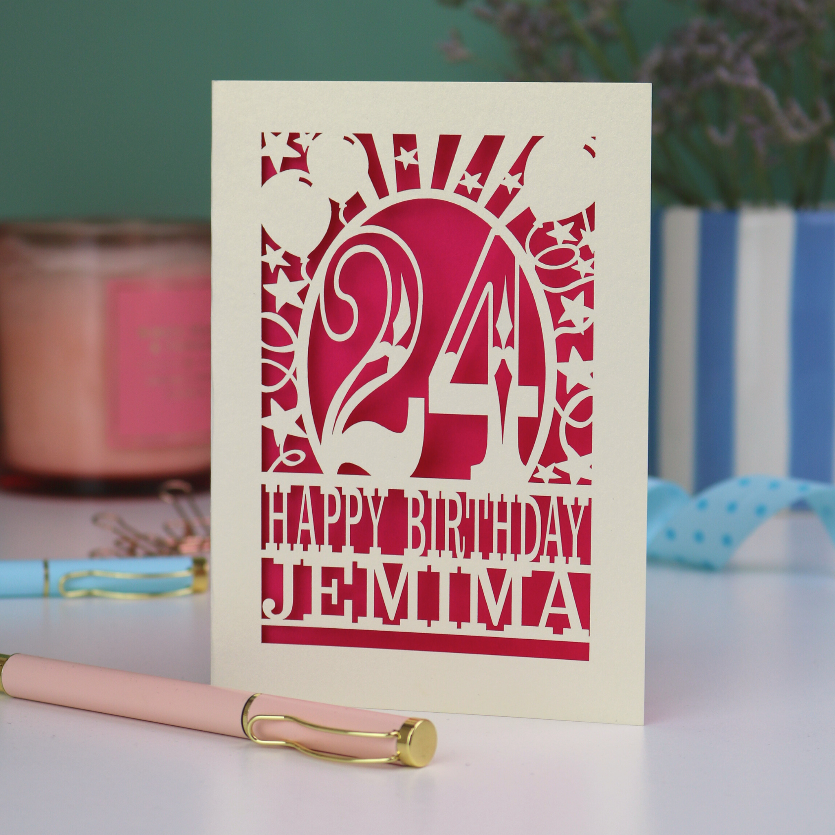 A cream laser cut birthday card on a white desk with stationery items. Card has the age 24 surrounded by stars and balloons, with "Happy Birthday Jemima" underneath in cut out text