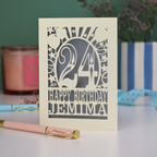 A cream and silver personalised laser cut birthday card on a white desk with stationery items. Card has the age 24 surrounded by stars and balloons, with "Happy Birthday Jemima" underneath in cut out text