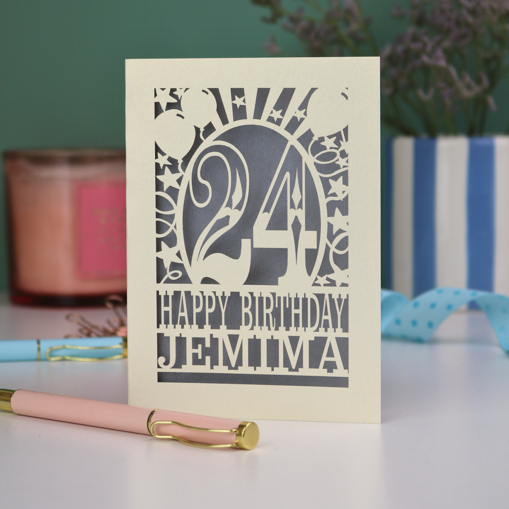 A cream and silver personalised laser cut birthday card on a white desk with stationery items. Card has the age 24 surrounded by stars and balloons, with "Happy Birthday Jemima" underneath in cut out text