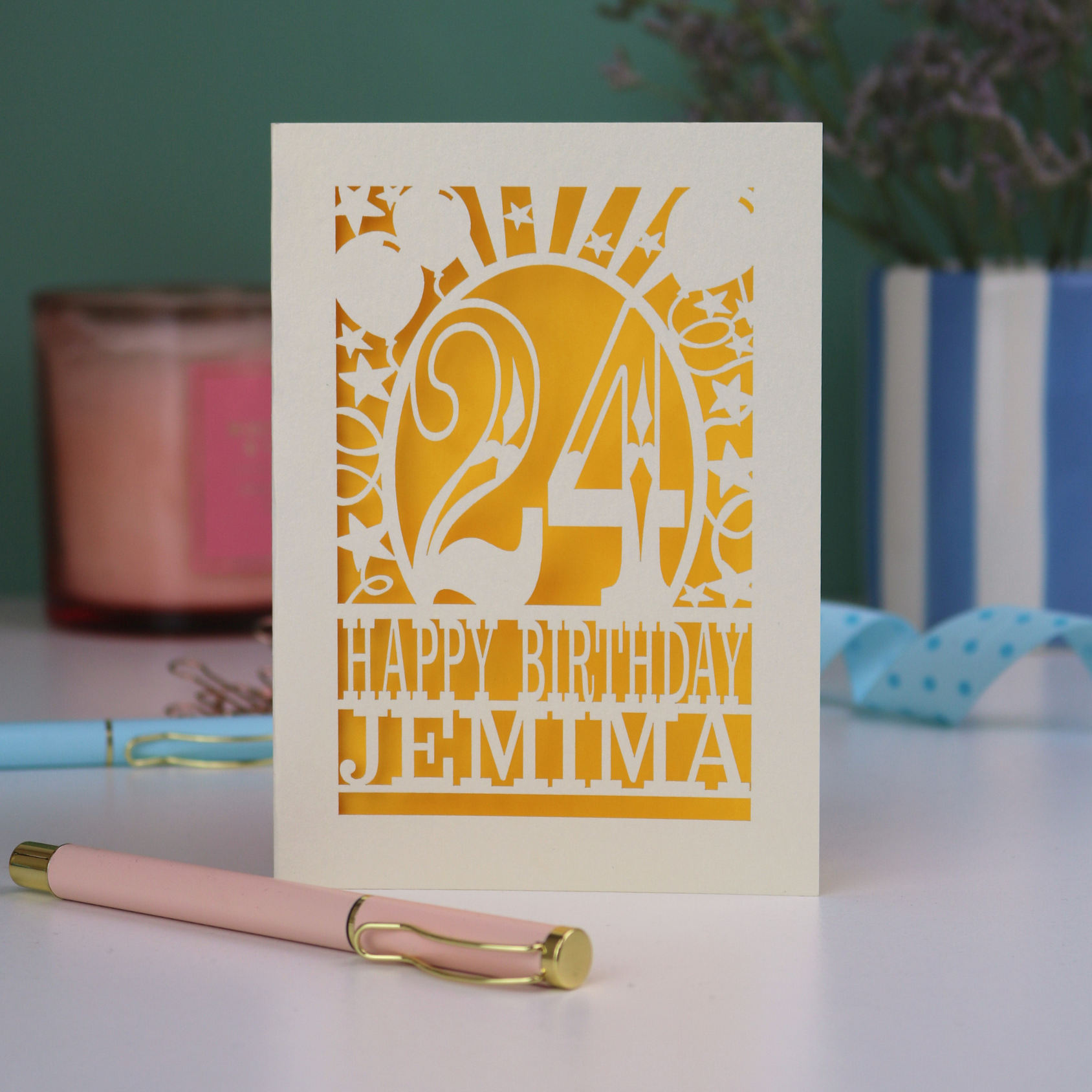 A cream and yellow laser cut birthday card on a white desk with stationery items. Card has the age 24 surrounded by stars and balloons, with "Happy Birthday Jemima" underneath in cut out text