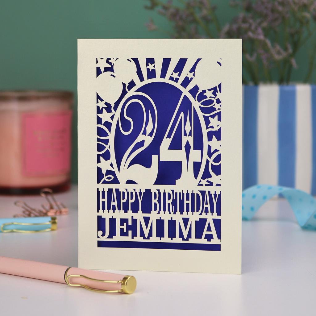 A cream laser cut birthday card on a white desk with stationery items. Card has the age 24 surrounded by stars and balloons, with "Happy Birthday Jemima" underneath in cut out text