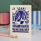 A cream laser cut birthday card on a white desk with stationery items. Card has the age 24 surrounded by stars and balloons, with "Happy Birthday Jemima" underneath in cut out text