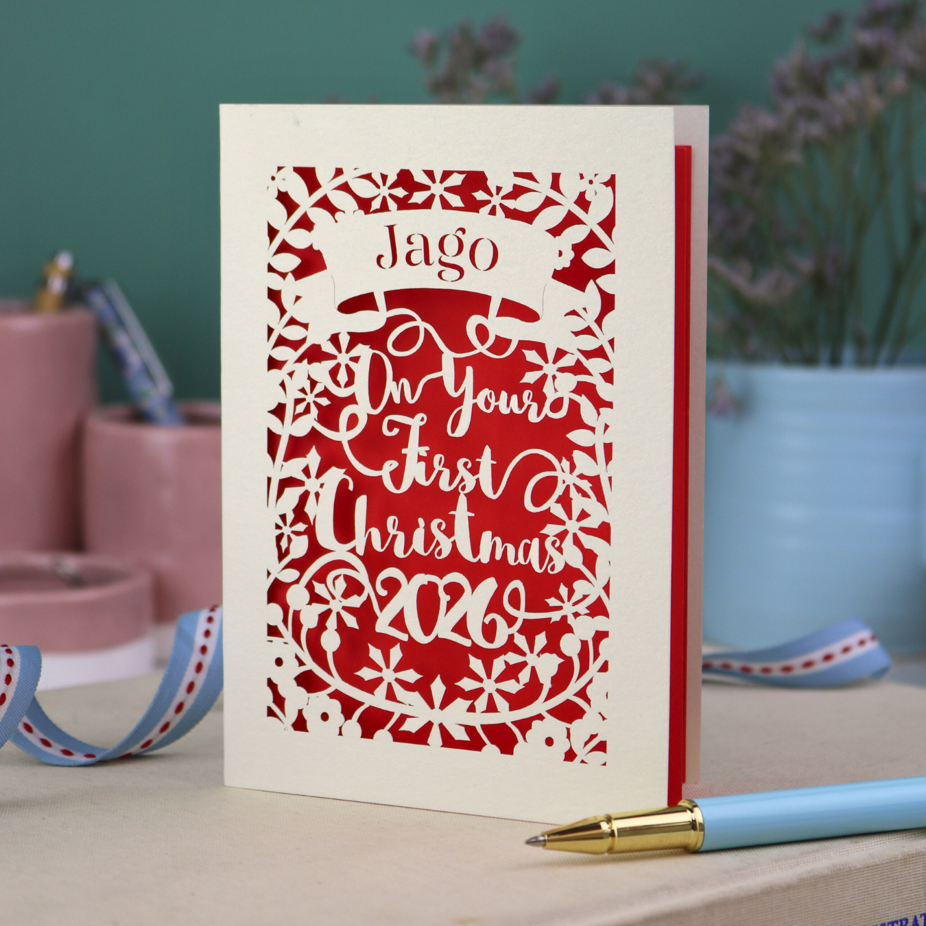 A cream and red laser cut baby's first Christmas card personalised with baby's name in a stencil font cut out writing on a banner, and the text "On your first Christmas" and the year 2026. 