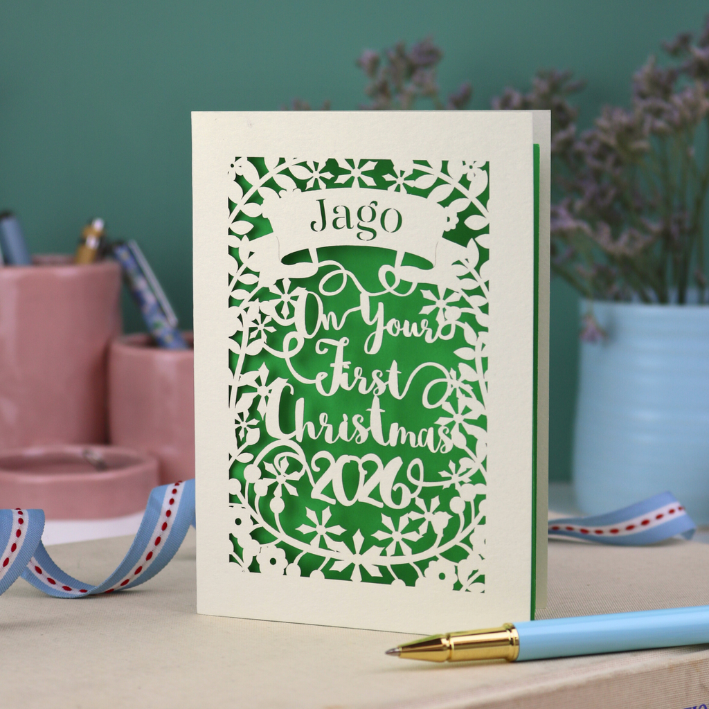 A laser cut baby's first Christmas card personalised with baby's name in a stencil font cut out writing on a banner, and the text "On your first Christmas" and the year 2026. 