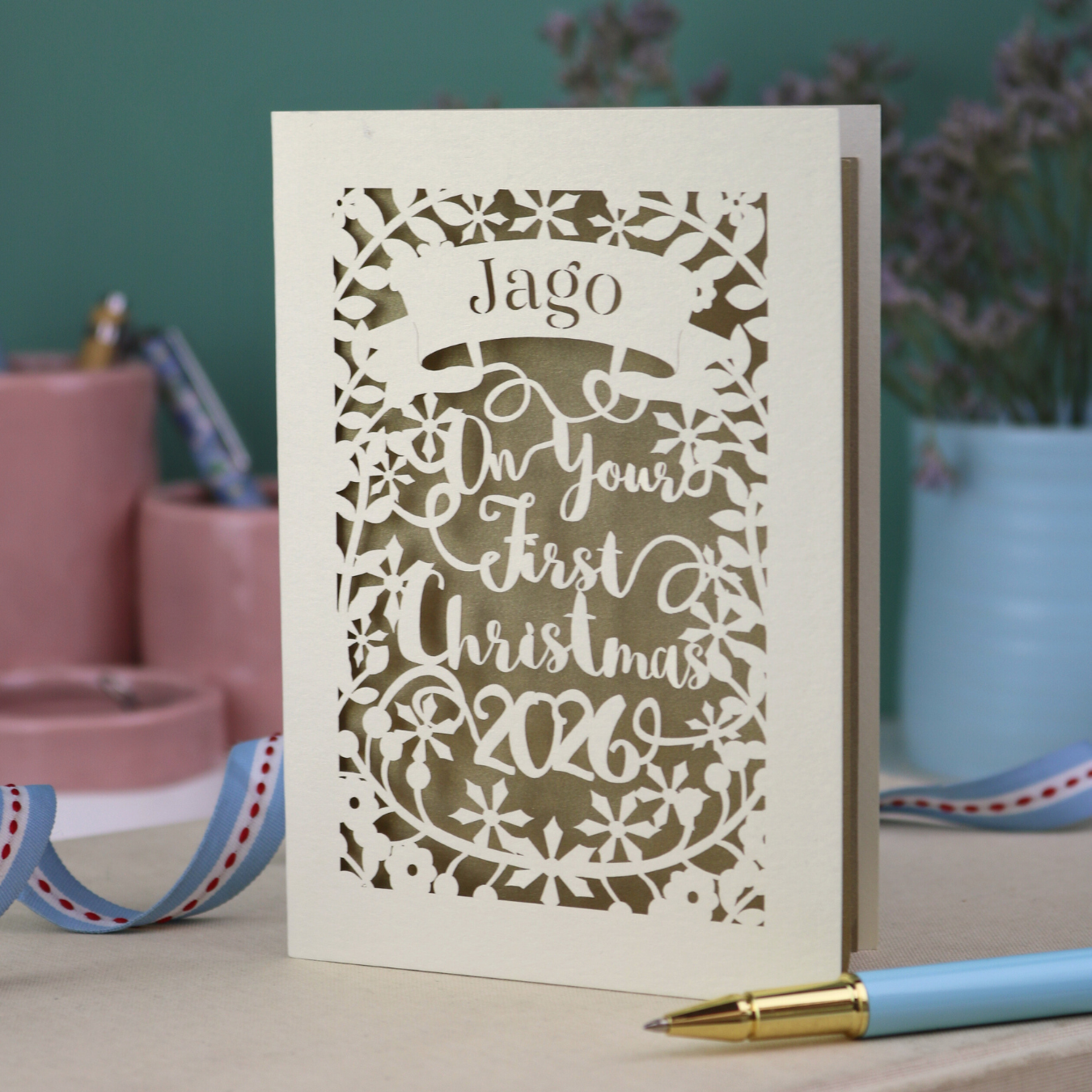 A laser cut baby's 1st Christmas card personalised with baby's name in a stencil font cut out writing on a banner, and the text "On your first Christmas" and the year 2026. 