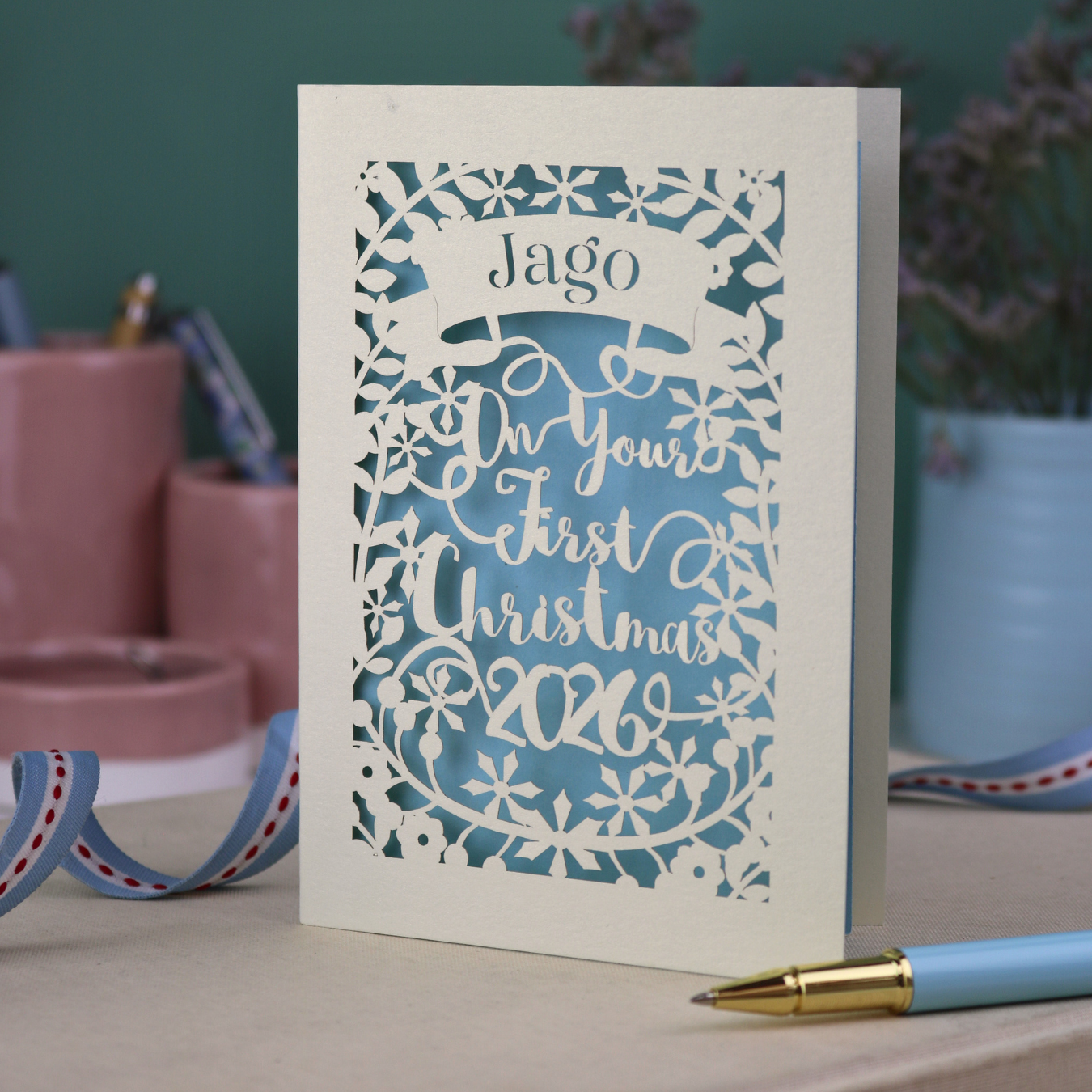 A laser cut baby's first Christmas card personalised with baby's name in a stencil font cut out writing on a banner, and the text "On your first Christmas" and the year 2026. 