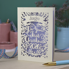 A paper cut baby's 1st Christmas card personalised with baby's name in a stencil font cut out writing on a banner, and the text "On your first Christmas" and the year 2026. 