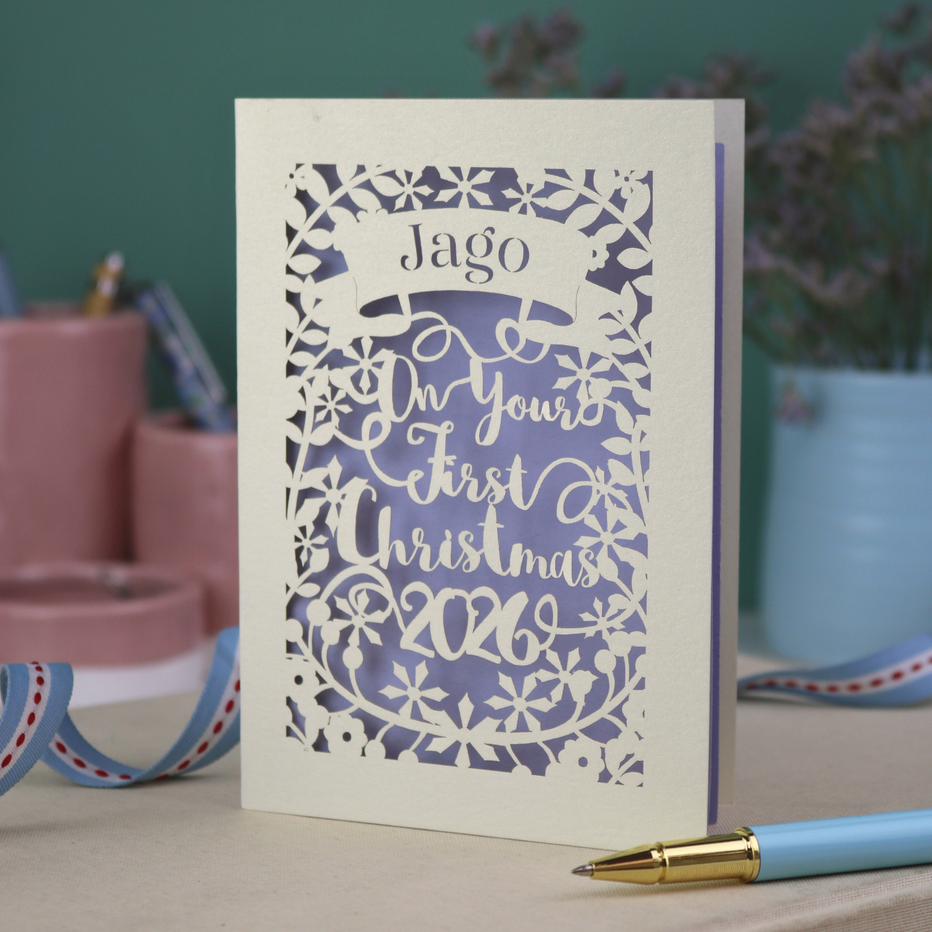 A paper cut baby's 1st Christmas card personalised with baby's name in a stencil font cut out writing on a banner, and the text "On your first Christmas" and the year 2026. 