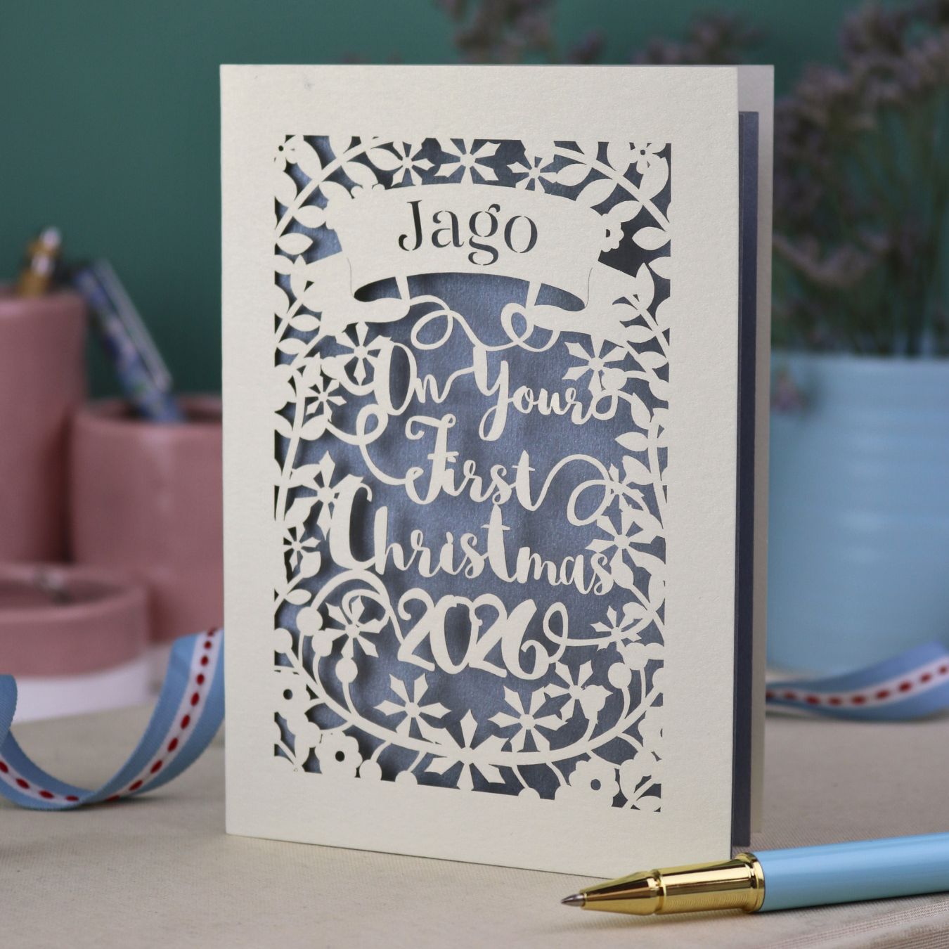 A laser cut keepsake baby's first Christmas card personalised with baby's name in a stencil font cut out writing on a banner, and the text "On your first Christmas" and the year 2026. 