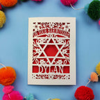 Papercut Bar Mitzvah Card with Star of David design and name 'Dylan' on a blue background with colourful pom-poms.
