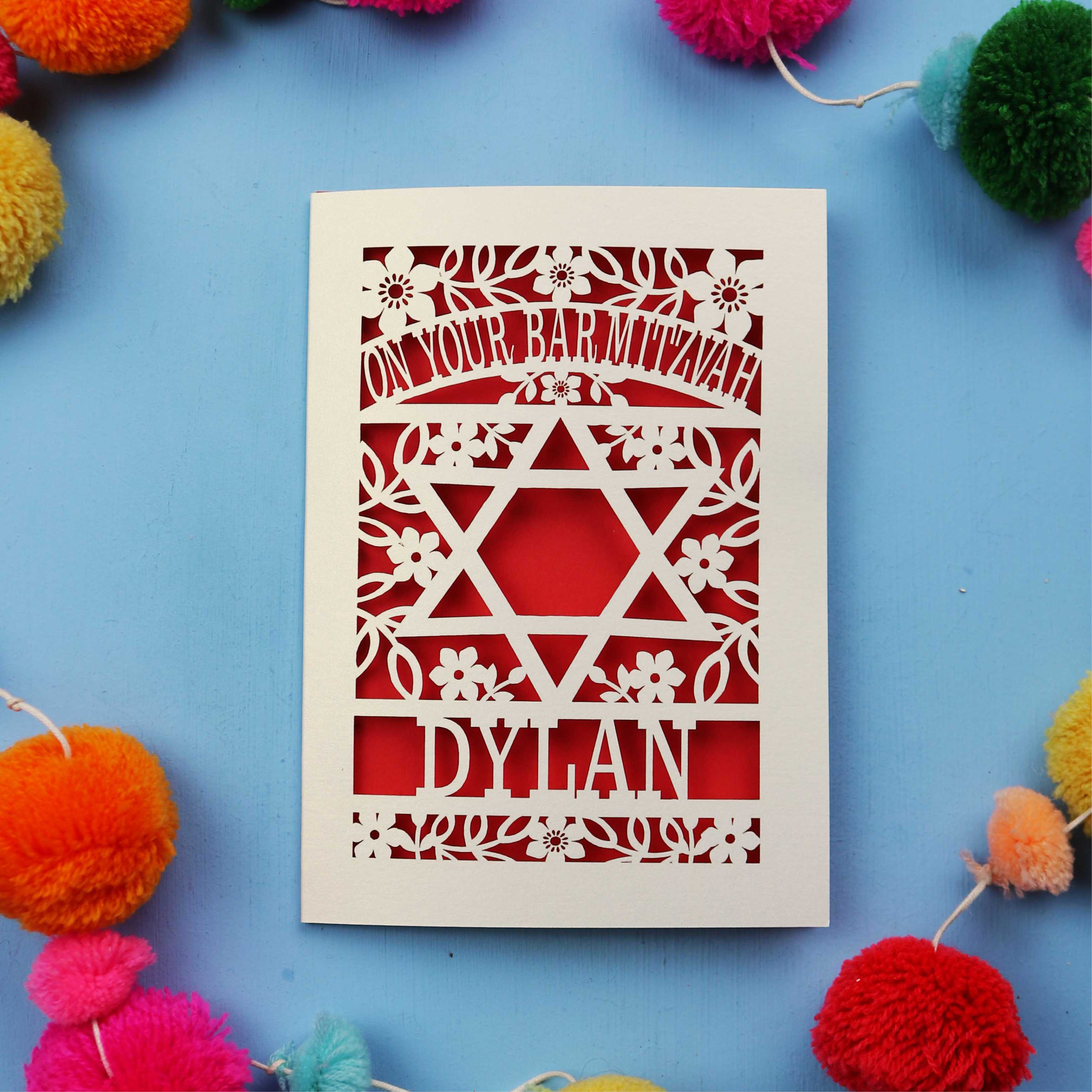 Papercut Bar Mitzvah Card with Star of David design and name 'Dylan' on a blue background with colourful pom-poms.