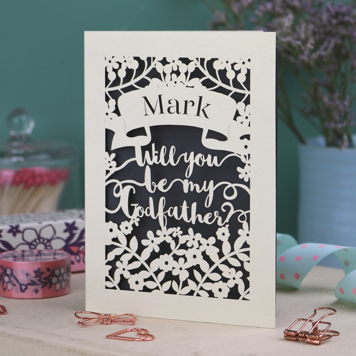 Laser-cut card asking 'Will you be my Godfather?' with decorative elements on a table.