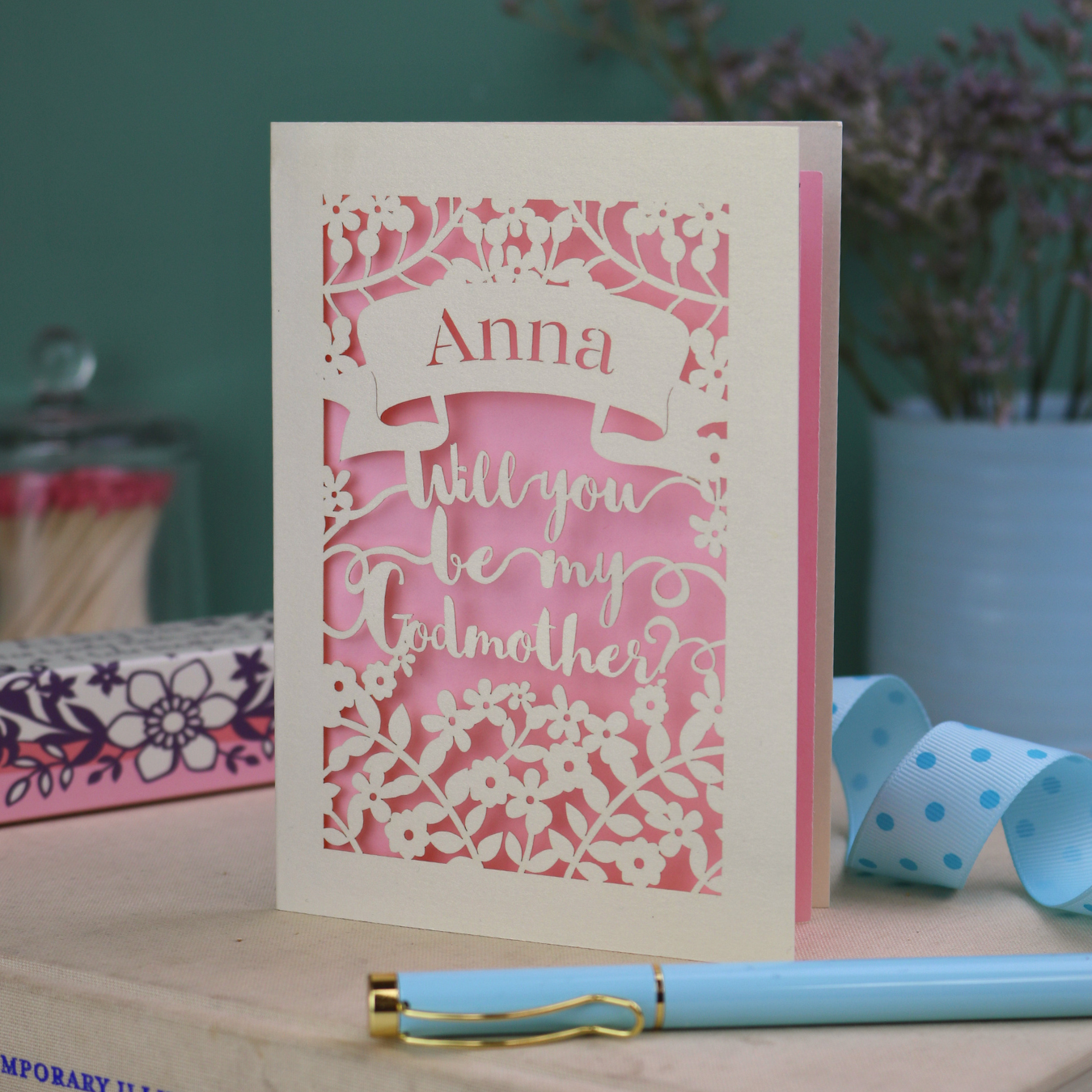 Personalised keepsake Godmother proposal card with 'Will you be my Godmother?' text and floral cut out border on a wooden surface with a pen and decorative items.