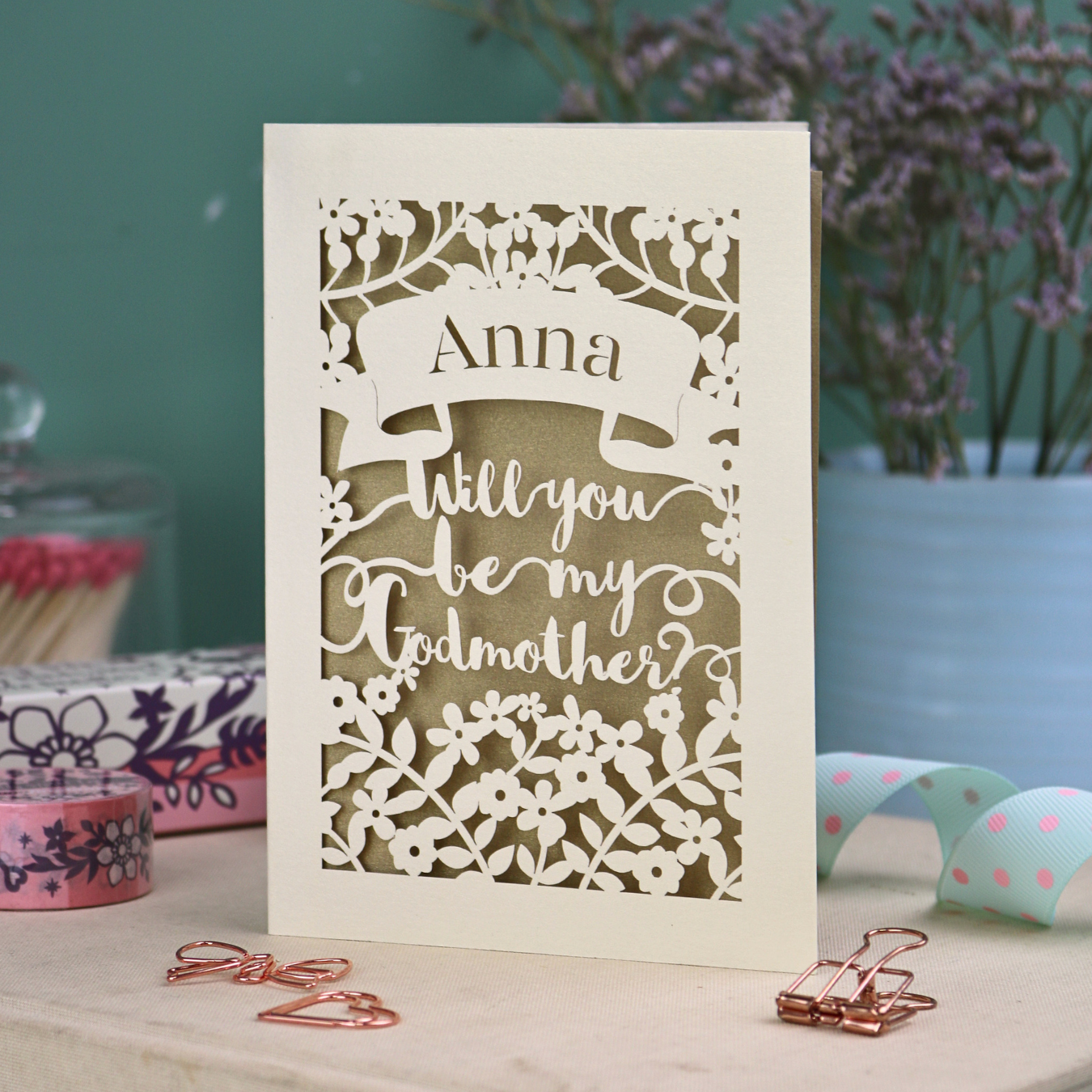 Laser-cut card asking 'Will you be my Godmother?' with decorative elements on a table.