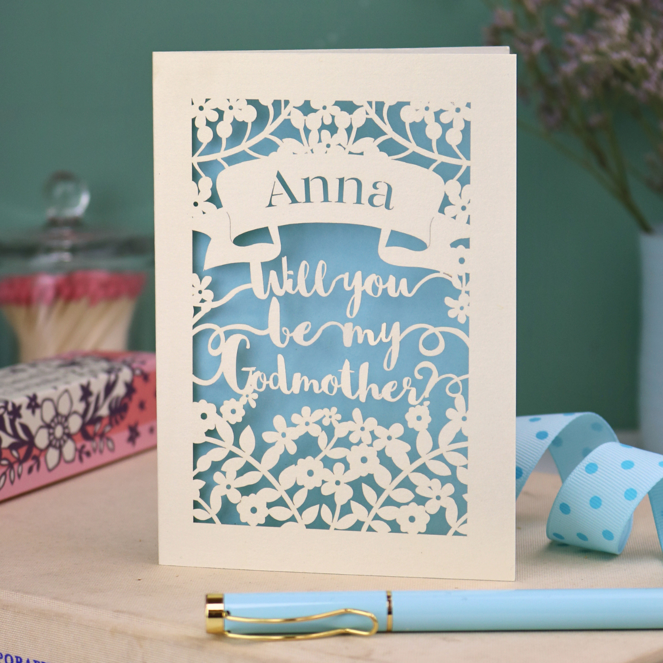 Personalised paper cut card with 'Will you be my Godmother?' text and floral cut out border on a wooden surface with a pen and decorative items.