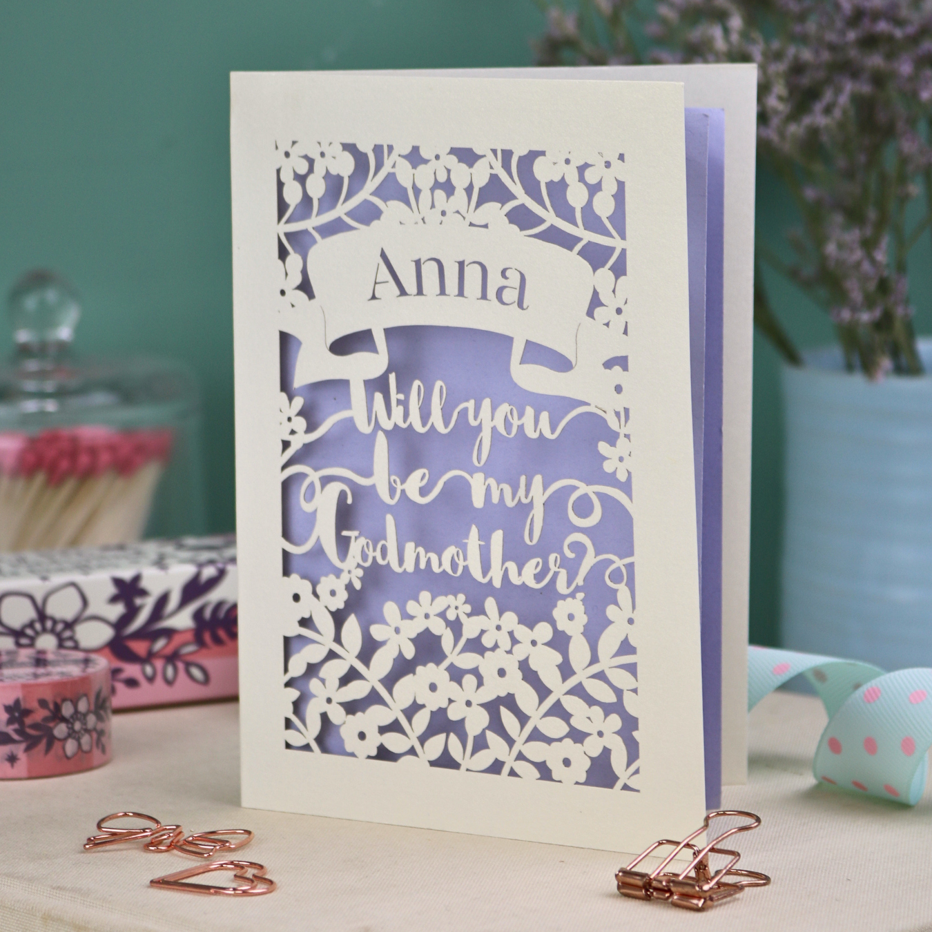 Personalised Godmother card with 'Will you be my Godmother?' text and floral cut out border on a wooden surface with a pen and decorative items.
