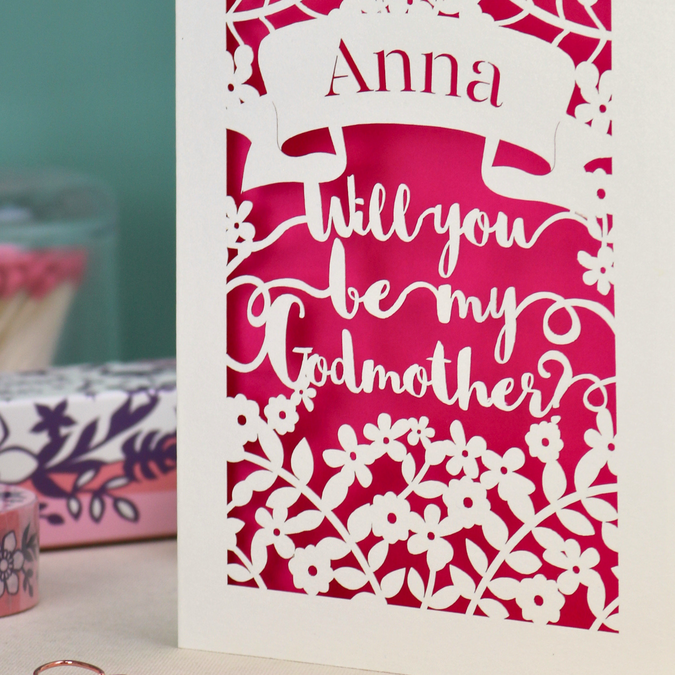 Personalised laser cut card with 'Will you be my Godmother?' text and floral cut out border on a wooden surface with a pen and decorative items.