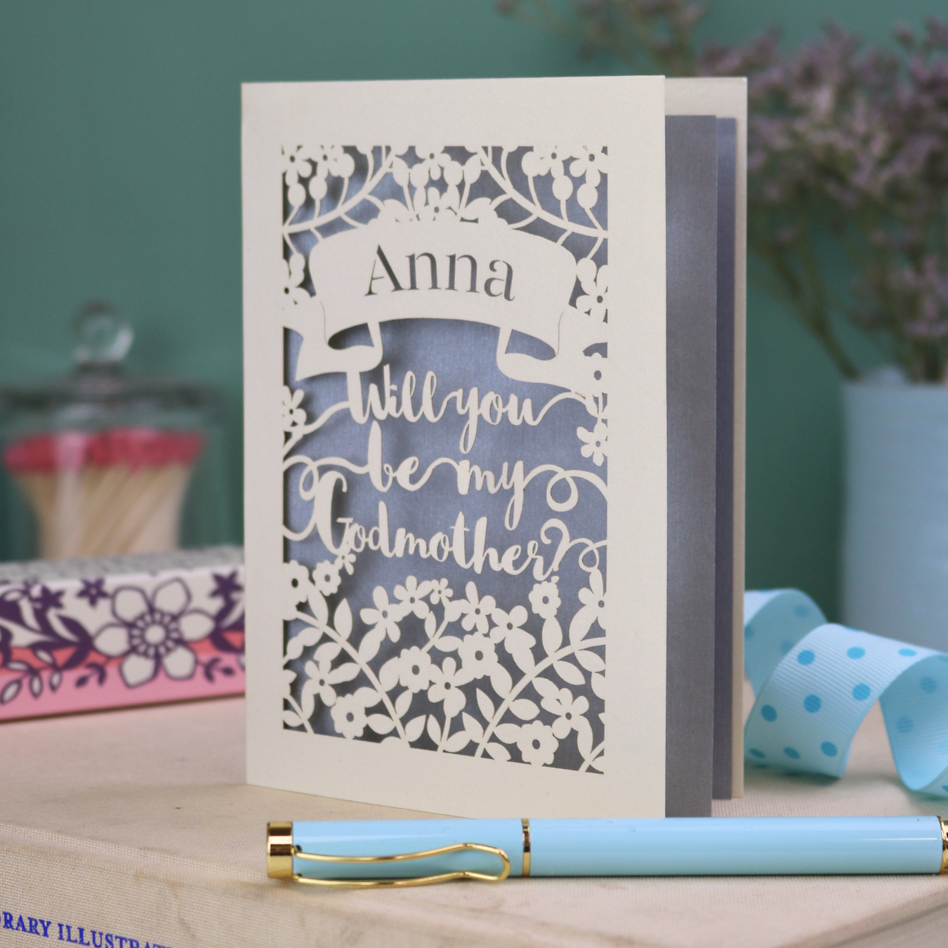 Personalised laser cut card with 'Will you be my Godmother?' text and floral cut out border on a wooden surface with a pen and decorative items.