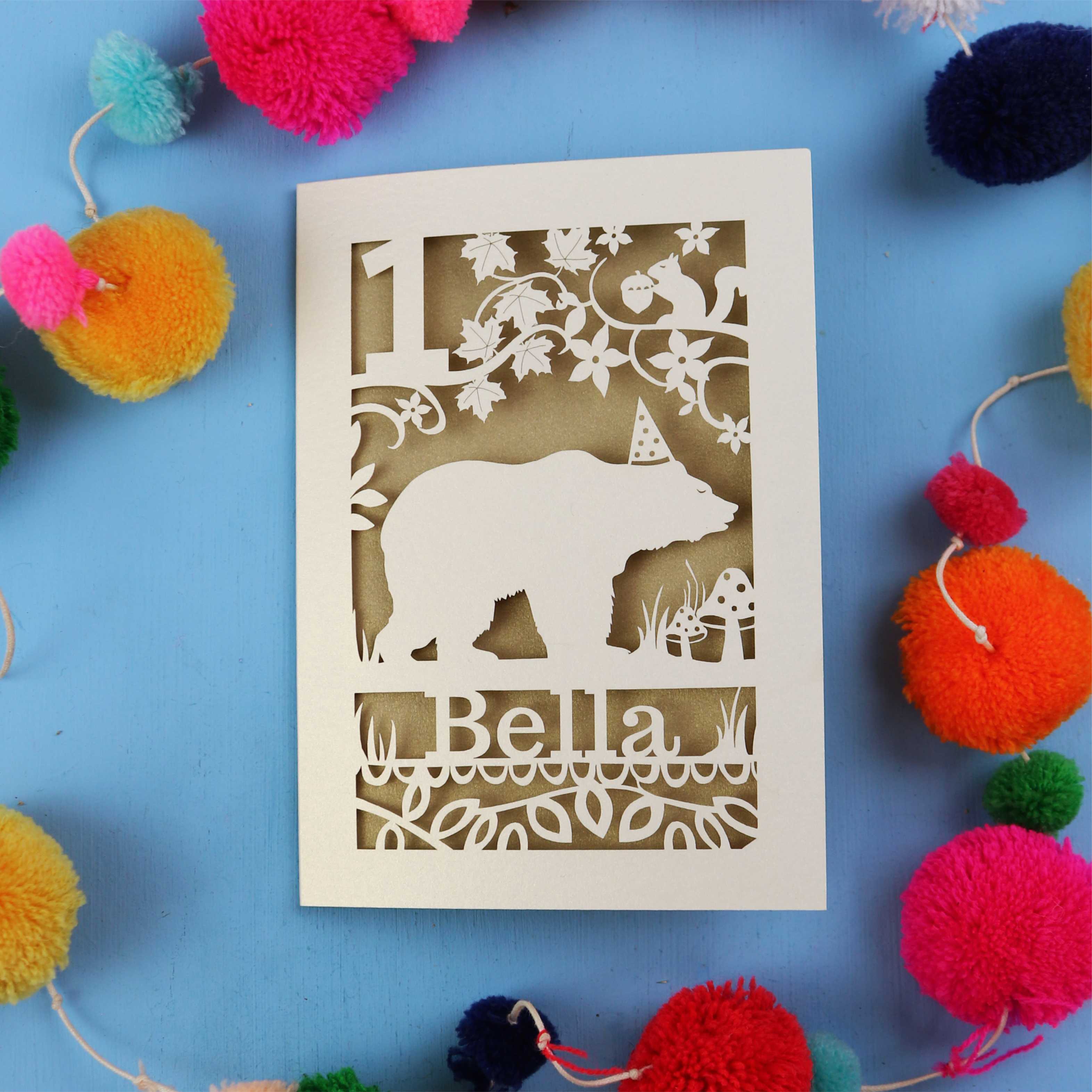 1st birthday Card with bear design and 'Bella' text on a blue background with colourful pom-poms.