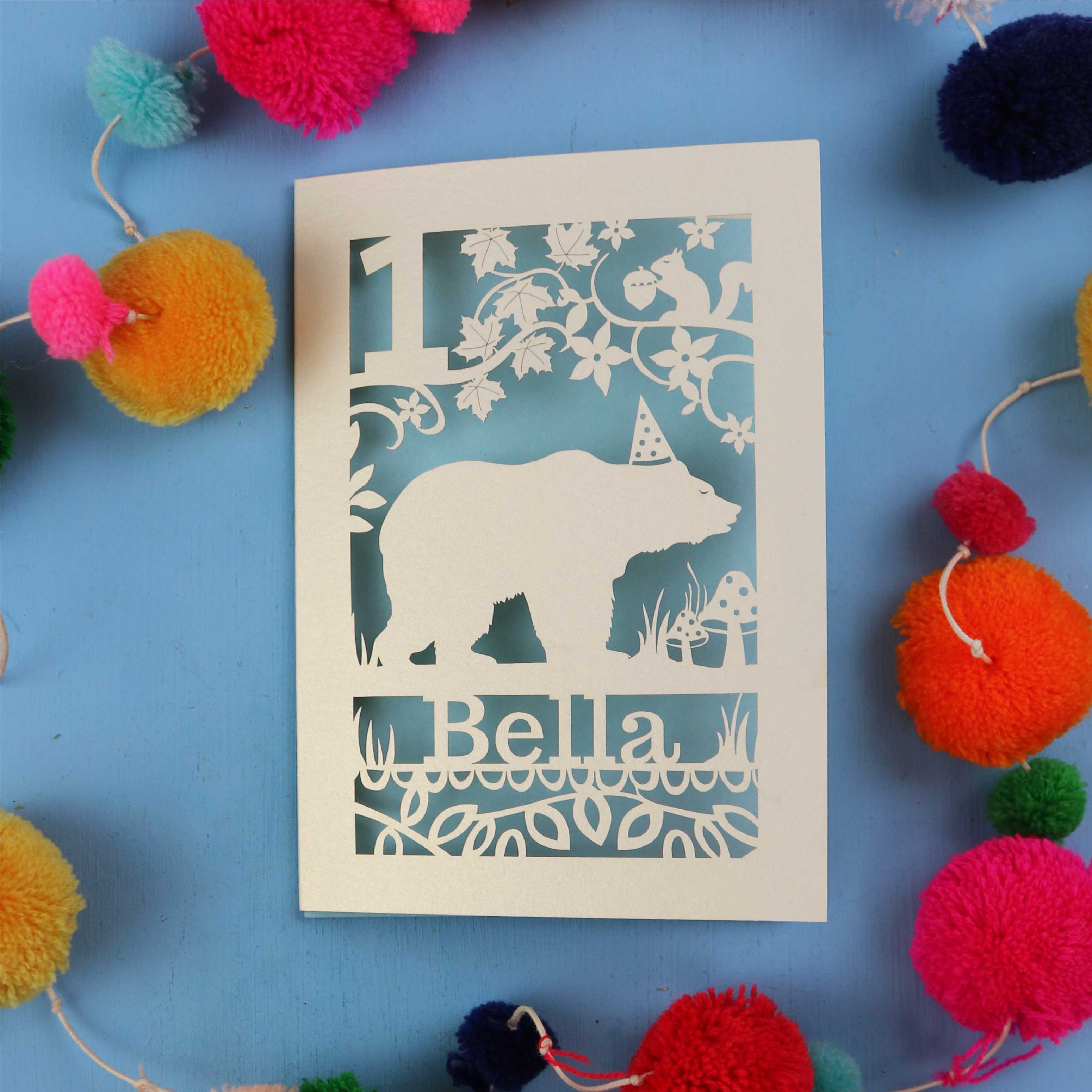laser cut 1st birthday Card with bear design and 'Bella' text on a blue background with colourful pom-poms.