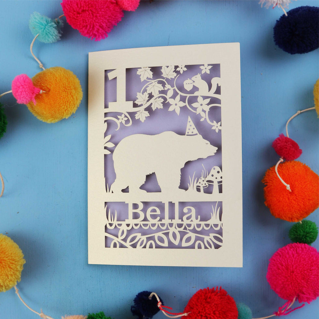 1st birthday Card with bear design and 'Bella' text on a blue background with colourful pom-poms.