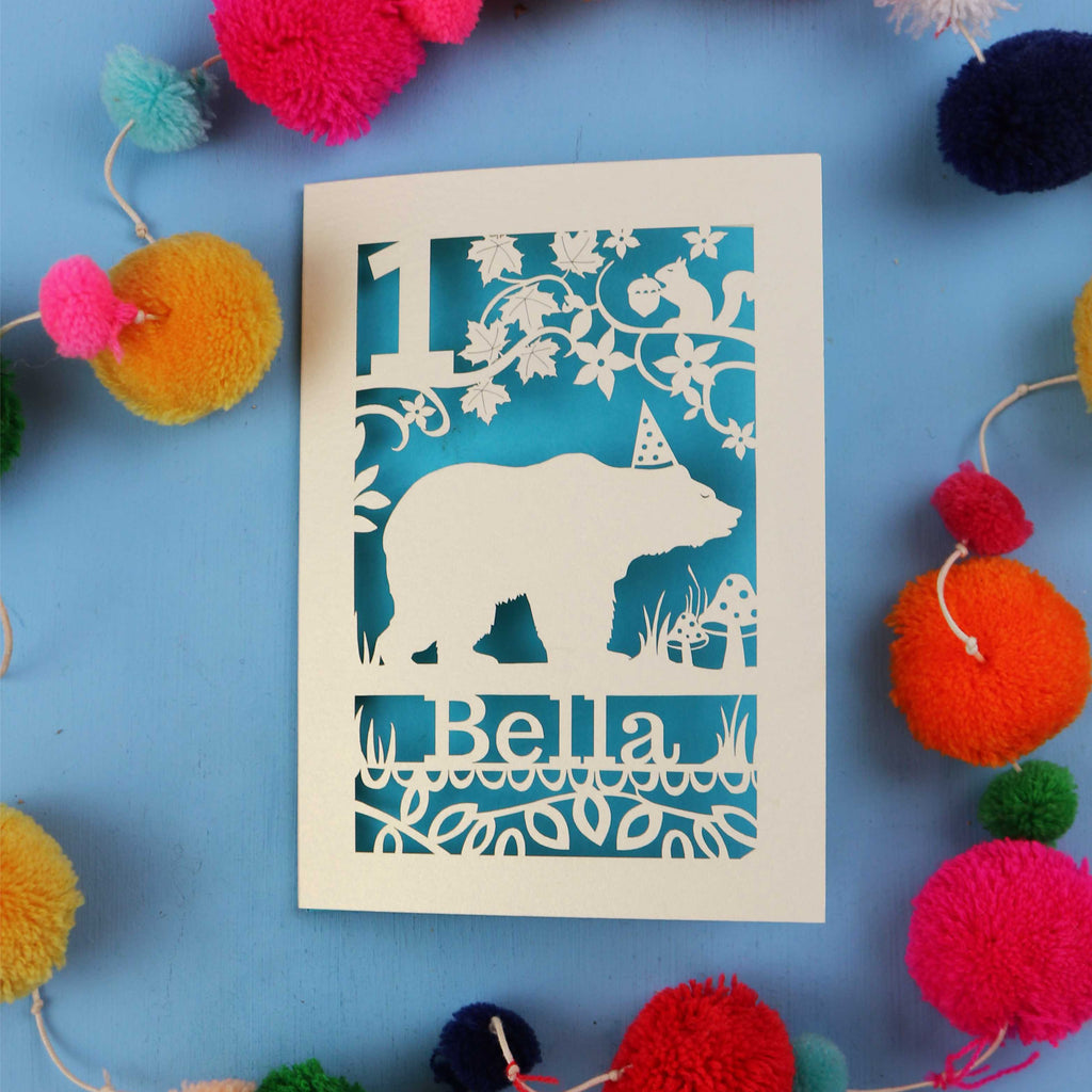 1st birthday Card with bear design and 'Bella' text on a blue background with colourful pom-poms.