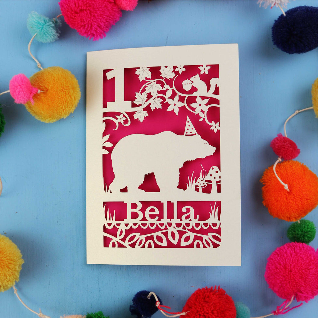 1st birthday Card with bear design and 'Bella' text on a blue background with colourful pom-poms.
