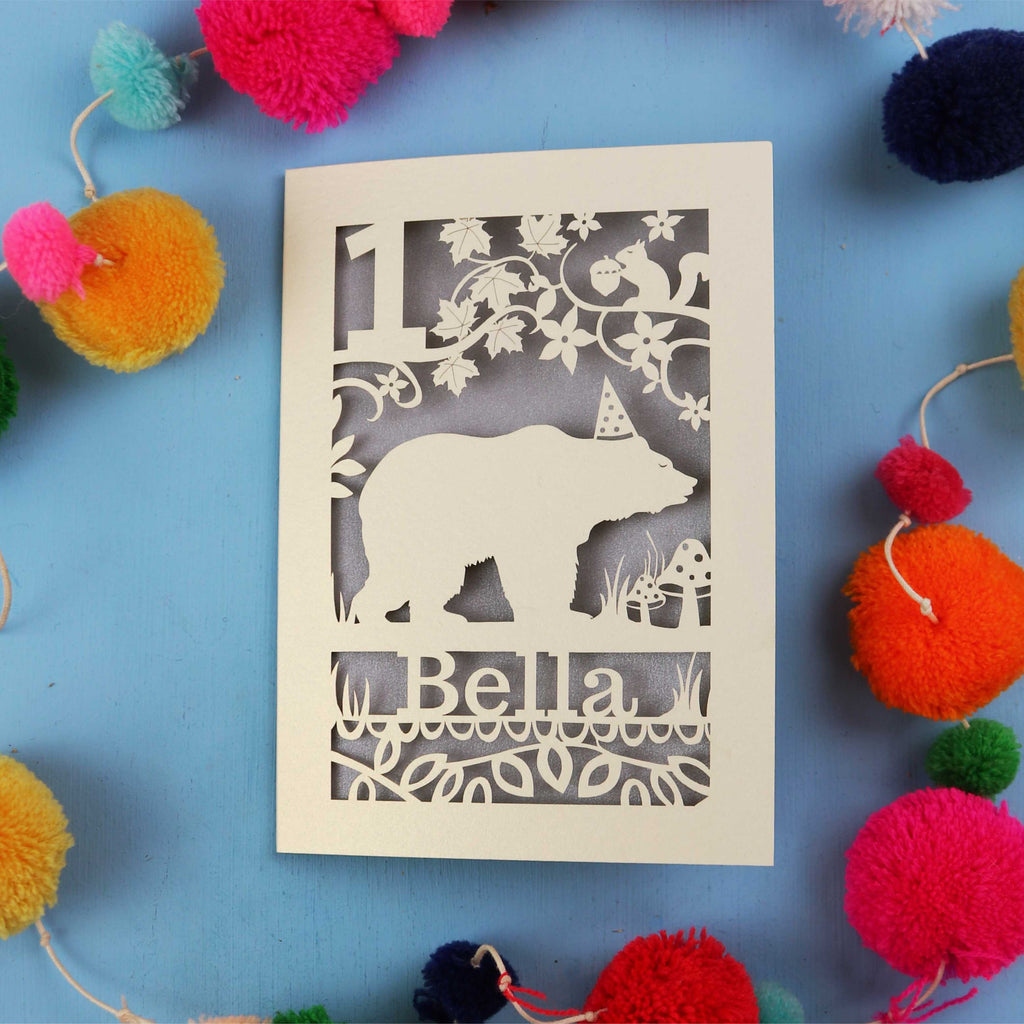 1st birthday Card with bear design and 'Bella' text on a blue background with colourful pom-poms.