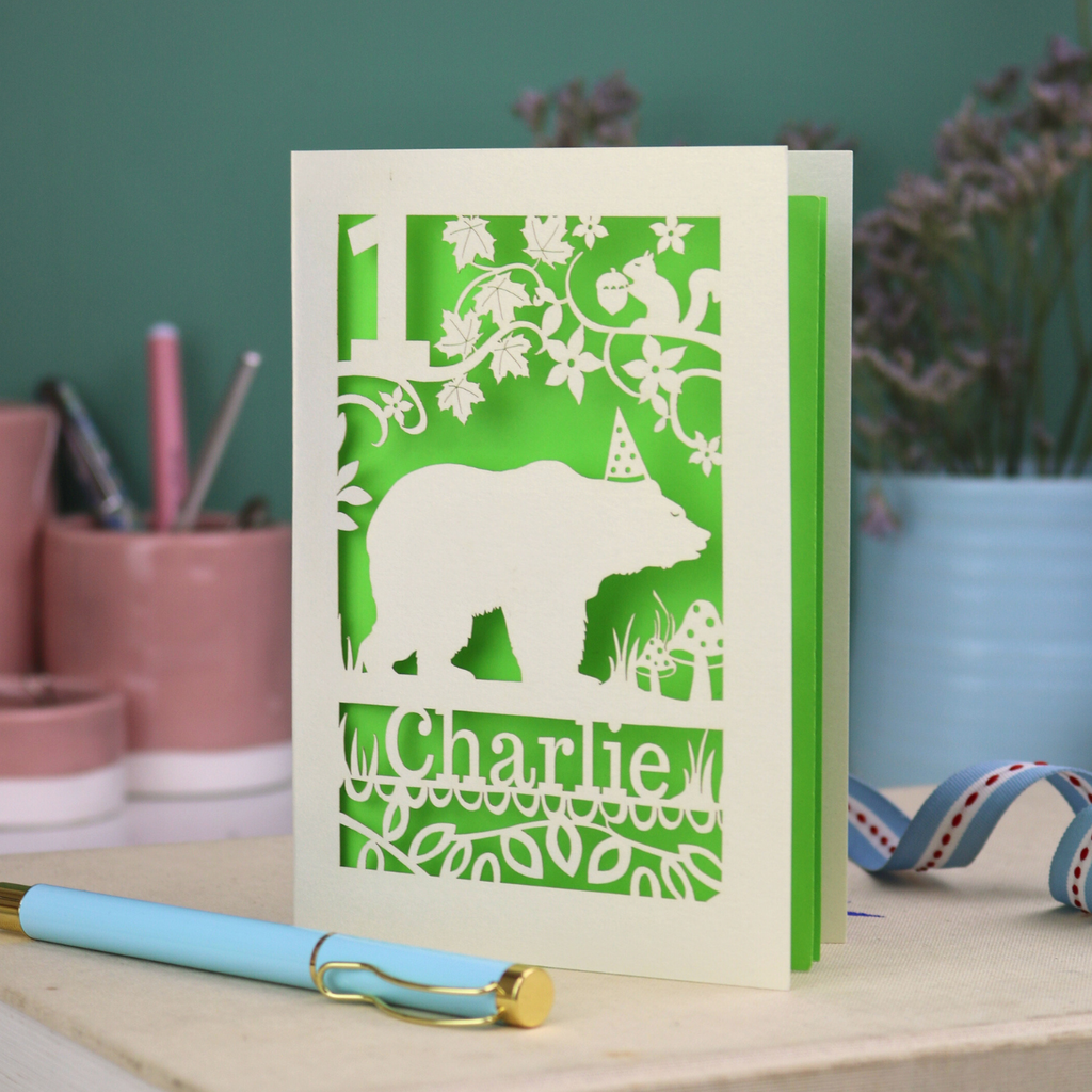 A laser cut birthday card personalised with an age and name, and featuring a cut out bear in a party hat with woodland border.