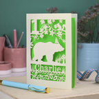 A laser cut birthday card personalised with an age and name, and featuring a cut out bear in a party hat with woodland border.