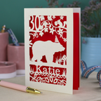 A laser cut birthday card personalised with an age and name, and featuring a cut out bear in a party hat with woodland border.