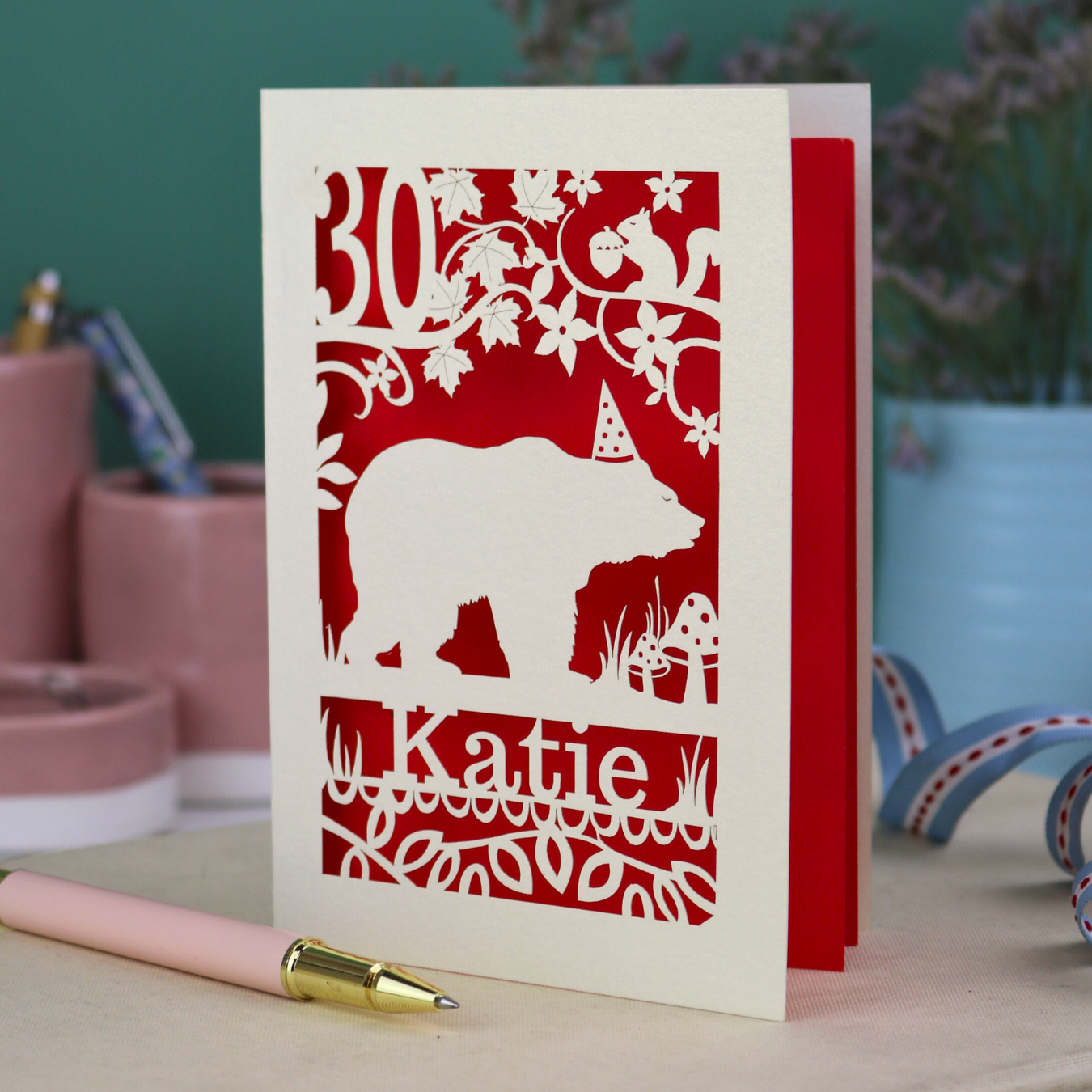 A laser cut birthday card personalised with an age and name, and featuring a cut out bear in a party hat with woodland border.