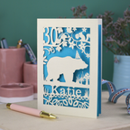 A laser cut birthday card personalised with an age and name, and featuring a cut out bear in a party hat with woodland border.